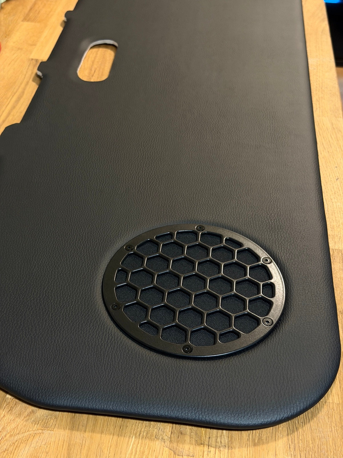 A black textured leather car interior trim panel with a circular honeycomb grille cutout, shown on a light wood floor.